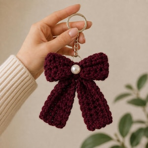 Puede incluir: Un llavero con forma de lazo tejido a ganchillo en color burdeos oscuro, con una perla blanca artificial en el centro. El lazo está unido a una anilla metálica plateada mediante una cadena corta, creando un accesorio texturizado de hilo rojo.