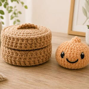 May include: A round, tan, crocheted lidded basket with a textured stitch pattern alongside a small, peach-colored, crocheted plush toy shaped like a dumpling with black embroidered eyes and a smiling mouth.