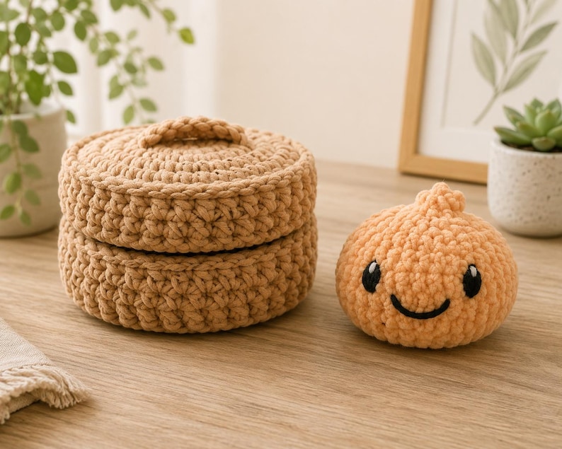 May include: A round, tan, crocheted lidded basket with a textured stitch pattern alongside a small, peach-colored, crocheted plush toy shaped like a dumpling with black embroidered eyes and a smiling mouth.
