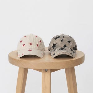 May include: Two baseball caps side by side. The left cap is off-white with small red embroidered stars. The right cap is a distressed grey with black printed stars and a frayed brim. Both feature a six-panel construction and a curved visor.