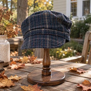 May include: A newsboy cap featuring a dark blue, black, and tan plaid pattern. The cap has a rounded crown, a structured brim, and a textured fabric construction. It is displayed on a wooden stand, showcasing the detailed weave of the material.