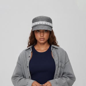 Op de afbeelding: Een grijze cadet-pet met een textuurrijke stoffen afwerking en een decoratieve witte kanten band rondom de kroon. De pet heeft een gestructureerde klep en een plat bovenstuk, wat zorgt voor een strakke en uniforme uitstraling van dit hoofddeksel.