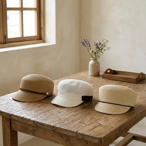 May include: Three structured caps with short brims, crafted from a woven straw-like material. The left cap is tan with a dark brown band, the center cap is white with a dark brown band, and the right cap is light beige with a dark brown band.