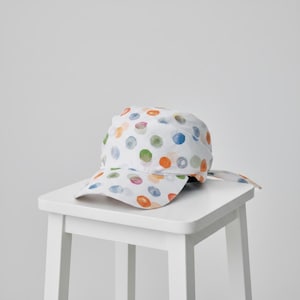 May include: A white baseball cap featuring a scattered pattern of watercolor-style polka dots in shades of orange, blue, green, and purple. The cap has a curved brim and fabric ties extending from the back, creating a soft, rounded silhouette.