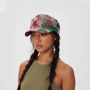 Puede incluir: Una gorra de estilo militar con un estampado floral multicolor en tonos rojos, verdes y blancos sobre un fondo rosa claro. El sombrero cuenta con una visera estructurada y un diseño de tela fruncida en la parte posterior para un ajuste ceñido.
