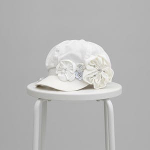 May include: A white newsboy cap featuring a structured brim and a rounded crown. The front is decorated with three fabric flowers of varying sizes, each accented with small clear beads and silver-toned centers, creating a layered floral arrangement.