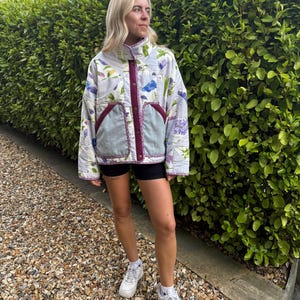 May include: A quilted jacket featuring a light grey base with a colourful floral print of blue and purple flowers and green leaves. The jacket has a high collar, a dark magenta front placket with metal snap buttons, and two large front pockets.
