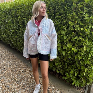 May include: A quilted jacket featuring a light blue and white floral pattern with pink trim, a high collar, and two front pockets. The jacket is worn over a red and white striped shirt and black shorts, paired with white trainers and white socks.