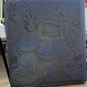 May include: A blue rectangular binder featuring a dark, laser-engraved illustration of the Pokemon Blastoise in the center, flanked by Gyarados on the left and Vaporeon on the right. The cover has a textured surface with a prominent central lock icon.