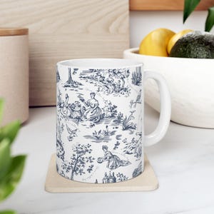 May include: A white ceramic mug featuring a dark blue toile de jouy pattern. The design displays intricate illustrations of people in historical attire, trees, birds, and landscape elements scattered across the surface of the cylindrical vessel.