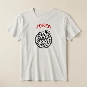 May include: White t-shirt with the word "JOKER" in red above a black circular design. The design features an eye and abstract shapes. The shirt is short-sleeved and has a crew neck.
