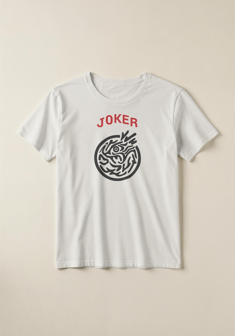 May include: White t-shirt with the word "JOKER" in red above a black circular design. The design features an eye and abstract shapes. The shirt is short-sleeved and has a crew neck.