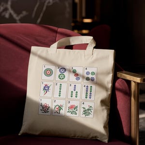 May include: Cream-colored tote bag featuring a colorful Mahjong tile design. The tiles display various suits and numbers, including circles, bamboos, and flowers. The bag has a simple, rectangular shape with two handles.