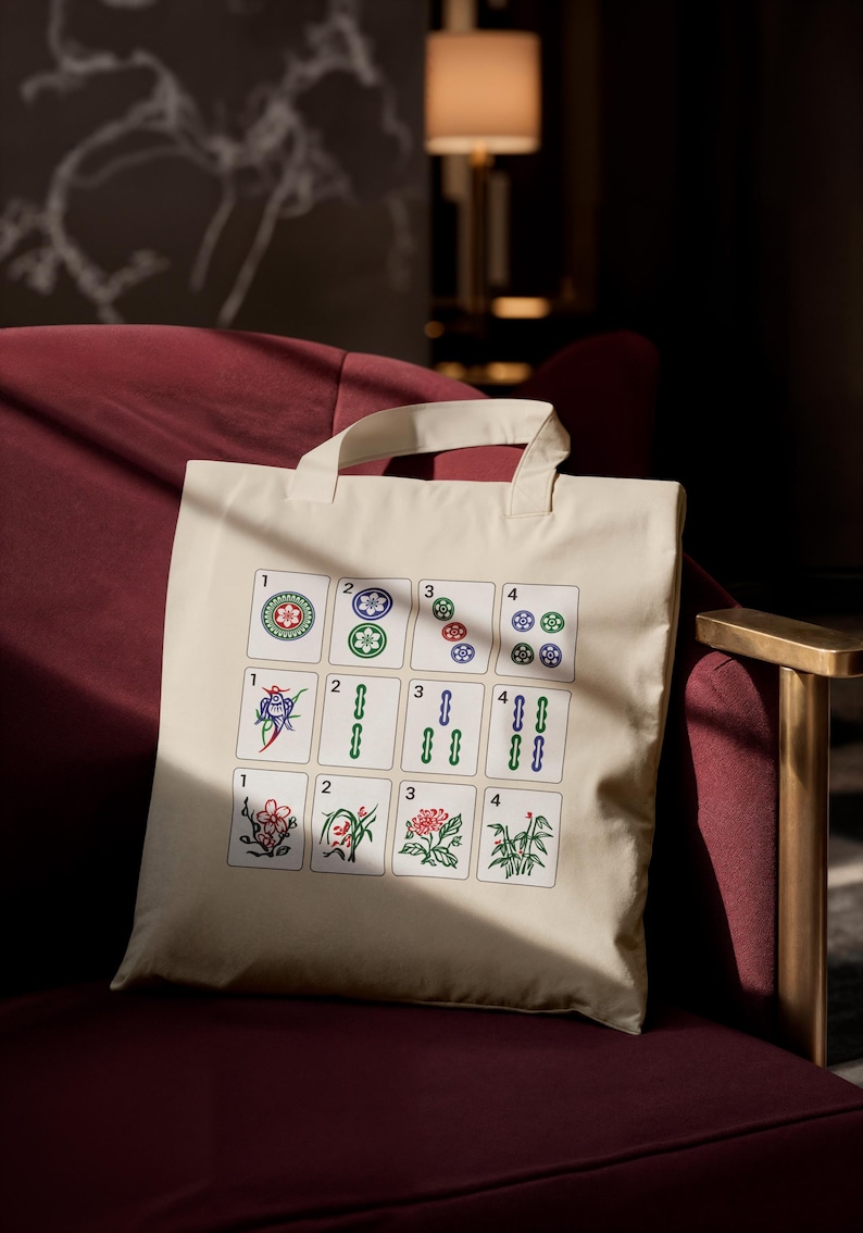 May include: Cream-colored tote bag featuring a colorful Mahjong tile design. The tiles display various suits and numbers, including circles, bamboos, and flowers. The bag has a simple, rectangular shape with two handles.