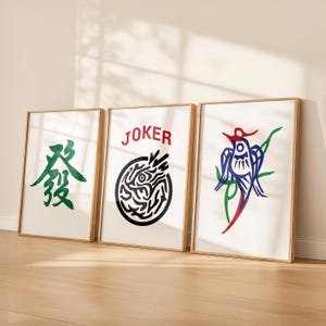 May include: Three framed art prints with gold-colored frames. The prints feature graphic designs: a green Chinese character, a black and white circular design with the word "JOKER" in red, and a blue, red, and green bird design.
