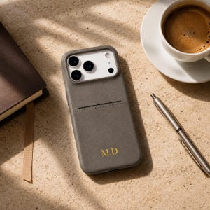 Peut inclure: Une coque de smartphone gris anthracite dotée d'une fente pour carte et des initiales M.D. dorées en relief en bas. La coque présente une découpe pour un système de triple objectif et une finition mate et lisse sur la surface arrière.