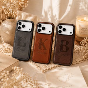May include: Three rectangular smartphone cases in black, tan, and dark brown leather, each featuring a large embossed initial letter and a cursive name underneath. The cases have a white camera module cutout and a smooth, matte finish.