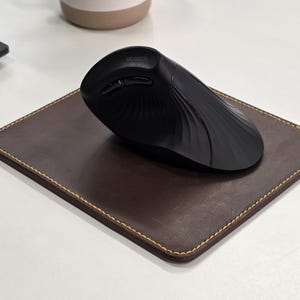 May include: A black ergonomic computer mouse rests on a brown leather mousepad with yellow stitching. The mouse has a curved design and several buttons. The mousepad is rectangular.