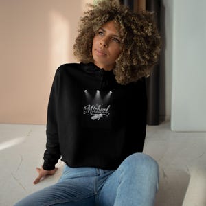 Puede incluir: Sudadera corta negra con el nombre "Michael" en fuente plateada, un gráfico de una mano y focos. La sudadera con capucha la lleva una persona con vaqueros azules.