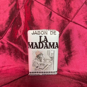 May include: A rectangular bar of soap wrapped in white paper with black text that reads JABON DE LA MADAMA ORIGINAL PRODUCTS. The label features a black and white line drawing of a person wearing a headscarf sitting at a table reading a book.