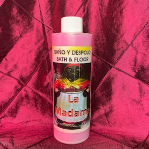 Peut inclure: Une bouteille en plastique cylindrique remplie de liquide rose, dotée d'un bouchon à vis blanc. L'étiquette porte le texte BAÑO Y DESPOJO BATH & FLOOR et La Madama au-dessus d'une illustration d'un personnage portant un châle jaune et un tablier blanc.