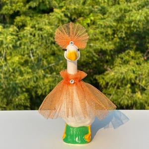 May include: A white ceramic goose figurine wearing an orange glittery tulle skirt and a matching orange tulle headpiece. The goose has a yellow beak, black eyes, and two small clear rhinestones centered on the headpiece and the skirt neckline.