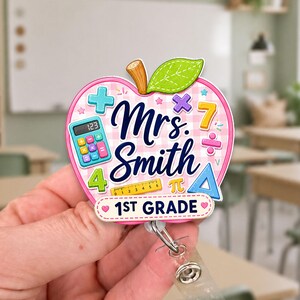 Peut inclure: Un porte-badge en forme de pomme avec un fond à carreaux roses, portant le texte Mrs. Smith et 1st Grade. Le design comprend des illustrations colorées d'une calculatrice, d'une règle, d'un triangle, d'un chiffre sept, d'un plus et d'une feuille verte.