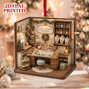 May include: A 2D flat printed hanging ornament depicting a detailed jewelry studio interior. The scene features a wooden desk with tools, a stool, shelves filled with jars of beads, jewelry displays, and a sign reading ELISA'S JEWELRY STUDIO.