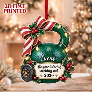 May include: A 2D flat printed kettlebell Christmas ornament in dark green with a red and white striped bow, holly berries, and string lights. It features the name Lucas, the text The year I started working out 2026, and a black snowflake emblem.