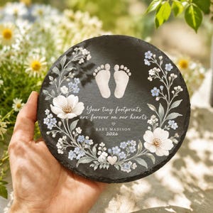 May include: A round black slate plaque featuring a white floral wreath design, a pair of baby footprints, and the text: Your tiny footprints are forever on our hearts, BABY MADISON, 2026. The plaque has a textured, natural stone edge.