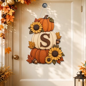 May include: A hanging wall decoration featuring a stack of three pumpkins in orange and cream colors. The center cream pumpkin displays a large dark brown letter S. The arrangement is accented with yellow sunflowers, brown acorns, and orange maple leaves.