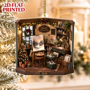 Può includere: Un ornamento natalizio stampato in 2D piatto che raffigura una accogliente stanza per dipingere con un paesaggio innevato su un cavalletto, un albero di Natale decorato, una scrivania con materiale artistico e la scritta Sarah's Painting Room e 2026.