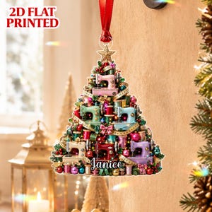 May include: A 2D flat printed Christmas tree ornament shaped like a pine tree, decorated with miniature sewing machines, colourful thread spools, and measuring tapes. The ornament features a red ribbon hanger and the name Janice in script text.