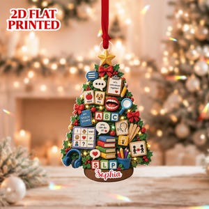 May include: A 2D flat printed Christmas tree ornament featuring speech therapy icons including an apple, dog, ABC blocks, books, a mouth, headphones, and a clipboard with the text 'let's communicate together'. The base is labeled with the name Sophia.