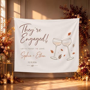 May include: A white fabric backdrop featuring the dark brown text They're Engaged!, Let's Toast to Love, Sophia + Ethan, and the date 10.10.2026. The design includes a line drawing of two clinking wine glasses surrounded by small, scattered autumn leaves.