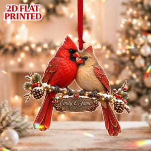 May include: A flat, 2D printed Christmas ornament featuring a bright red cardinal and a tan cardinal perched on a snowy branch with pinecones and red berries. A wooden-style plaque hangs from the branch with the text Emily & James 12.14.2026.