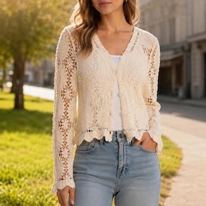 Könnte beinhalten: Eine cremefarbene, langärmelige Häkelstrickjacke mit V-Ausschnitt, durchgehender Knopfleiste und komplizierten, blumenartigen Lochmustern im gesamten Strickgewebe. Das Kleidungsstück wird über einem schlichten weißen Oberteil und einer hellblauen Jeans getragen.
