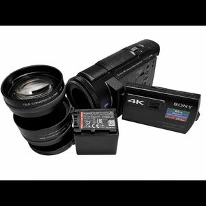 Könnte beinhalten: Ein schwarzer Sony 4K Handycam Camcorder mit 64 GB internem Speicher, einem rechteckigen NP-FV100A Akku und zwei runden Objektivaufsätzen mit der Beschriftung Tele Conversion Lens und Wide Conversion Lens, alles vor weißem Hintergrund.