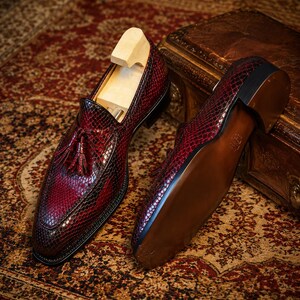 May include: A pair of burgundy leather loafers with a snakeskin pattern and tassel detailing. The shoes are displayed with wooden shoe trees inside, showcasing their shape and construction. The soles are brown with a black heel.