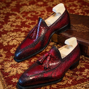 May include: A pair of red and black leather loafers with a snakeskin pattern. The shoes have a tassel detail and are displayed on a patterned rug. Shoe trees are inserted in the shoes.