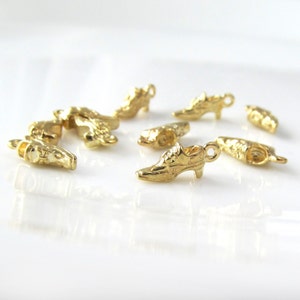 May include: A collection of gold-coloured shoe charms, some with an intricate design, others with a simple shape. The charms are scattered on a white surface.