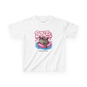 Peut inclure: T-shirt blanc avec une grenouille de dessin animé portant des lunettes de soleil et flottant dans une bouée rose. La grenouille a un motif léopard. Le mot "Boujee" est au-dessus de la grenouille en script rose, avec le texte "playground dept." en dessous.