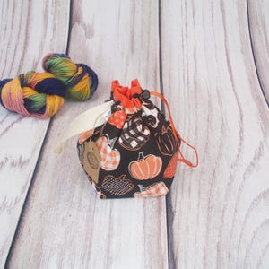 May include: A black drawstring bag with a pumpkin print. The bag is made of fabric and has a white drawstring. The bag is sitting on a white wooden surface.