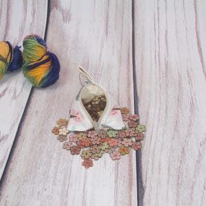 May include: A white fabric pouch with a floral print and a zipper closure. The pouch is filled with small, colorful wooden buttons. Two balls of yarn in shades of blue, yellow, and orange are in the background.