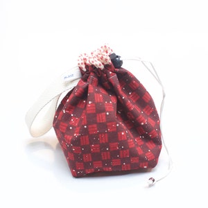 Peut inclure: Un petit sac à cordon avec un motif à carreaux bordeaux et rouge et des mouchetures blanches. Le haut est froncé avec un cordon blanc et un bouton noir. Une sangle blanche avec le texte "m.knitt" est attachée sur le côté.