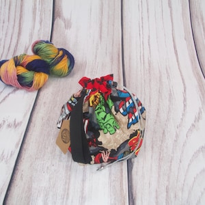 May include: A colorful drawstring bag with a superhero theme. The bag is made of fabric and has a black drawstring. The bag is sitting on a white wooden surface.