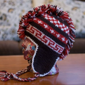 May include: A brown, red, and white knitted hat with a long, brown ear flap. The hat has a mohawk-style design with a white pom-pom on top.