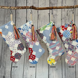 May include: Four patchwork fabric Christmas stockings with red, blue, and white patterns and tassels. The stockings are hanging from a branch on a white wood background. The stockings are labeled 004-001, 004-002, 004-003, and 004-004.