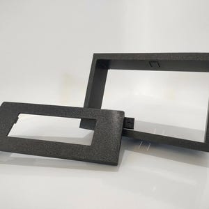 May include: Two black rectangular frames with a textured finish. The larger frame is positioned upright, while the smaller frame lies flat in front, featuring a small protruding tab on the left side and a rectangular cutout in the centre.