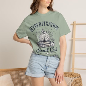 May include: A sage green short-sleeve t-shirt featuring a dark graphic print with the text "HYPERFIXATION Social Club" and "THE RABBIT HOLE". The design includes a brain topped with a butterfly, resting on books, next to a mug and flowers.